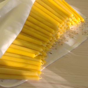 Op de afbeelding: Een bundel gele bijenwaskaarsen zijn gestapeld op een witte doek. De kaarsen zijn in een waaiervorm gerangschikt, met de toppen van de kaarsen die naar de rechterkant van de afbeelding wijzen.