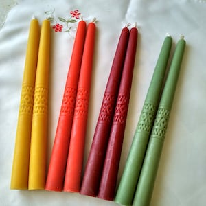 Op de afbeelding: Zes tapervormige kaarsen in verschillende kleuren, waaronder geel, rood, bordeauxrood en groen. De kaarsen hebben een decoratief patroon en zijn in een rij op een wit oppervlak gerangschikt.