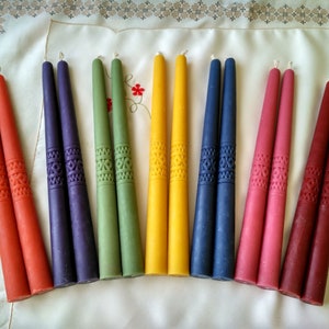 Op de afbeelding: Een set van 10 tapers in verschillende kleuren, waaronder oranje, paars, groen, geel, blauw en rood. De kaarsen hebben een decoratief patroon en zijn in een rij op een witte tafelkleed gelegd.