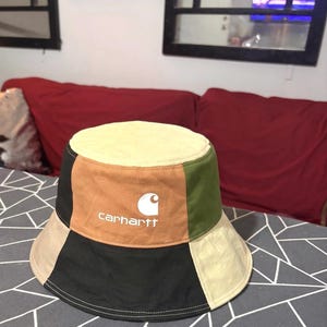 Puede incluir: Un sombrero de pescador con bloques de color en beige, marrón, verde oliva y negro. El panel frontal marrón muestra el logotipo y el texto de Carhartt en blanco. El sombrero tiene una copa plana y un ala inclinada con costuras blancas visibles.