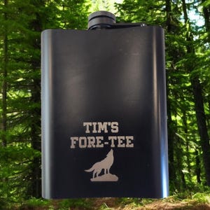 Pu&ograve; includere: Fiaschetta nera con la scritta "TIM'S FORE-TEE" e una grafica di un lupo che ulula. La fiaschetta ha un tappo nero ed &egrave; ambientata su uno sfondo di alberi verdi. Un regalo personalizzato per un appassionato di golf.