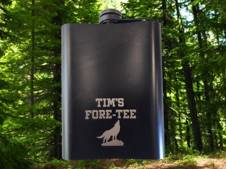Pu&ograve; includere: Fiaschetta nera con la scritta "TIM'S FORE-TEE" e una grafica di un lupo che ulula. La fiaschetta ha un tappo nero ed &egrave; ambientata su uno sfondo di alberi verdi. Un regalo personalizzato per un appassionato di golf.