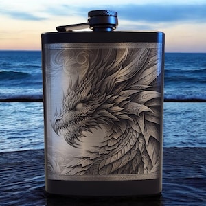 May include: A black stainless steel flask with a detailed dragon illustration on the front. The dragon is silver and gray, with intricate scales and a fierce expression. The flask has a black cap and hinge.