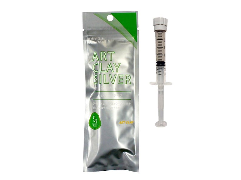 May include: A silver-colored foil packet labeled ART CLAY SILVER with a green accent and a 5g weight indicator, accompanied by a clear plastic syringe applicator with a white cap and black measurement markings along the side of the barrel.