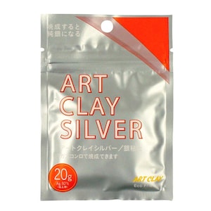 May include: A silver-colored, rectangular foil pouch containing Art Clay Silver. The front features the text ART CLAY SILVER in red, a circular 20g label, and Japanese characters. The top right corner has a bright orange triangular accent.