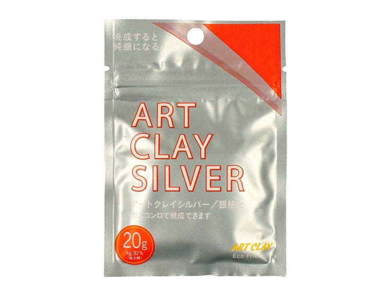 May include: A silver-colored, rectangular foil pouch containing Art Clay Silver. The front features the text ART CLAY SILVER in red, a circular 20g label, and Japanese characters. The top right corner has a bright orange triangular accent.