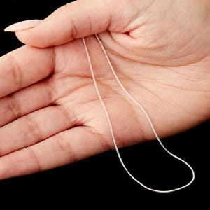 May include: A fine silver chain necklace, displayed against a dark backdrop. The chain is slender, composed of tiny, linked segments. The necklace has a simple, elegant design, suitable for daily wear or special events.