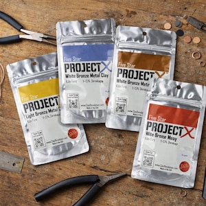 May include: Four silver pouches of Five Star PROJECT metal clay with coloured labels: yellow, blue, brown, and red. The text on the pouches includes "Light Bronze", "White Bronze", and "Wite Bronze". Tools and metal pieces are scattered around.