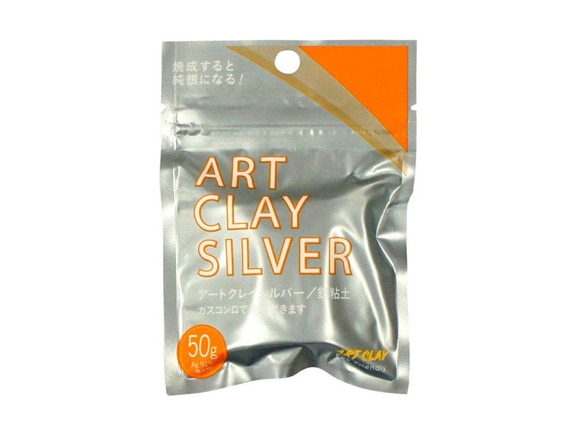 May include: A silver-colored, rectangular, flexible pouch containing 50g of Art Clay Silver. The packaging features the text ART CLAY SILVER in large letters, an orange corner accent, and Japanese characters printed on the front surface.