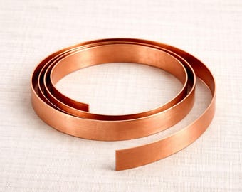 May include: A coiled length of copper strip, displaying a polished, reflective surface. The strip is unwound, showing its flat, rectangular form. The copper has a warm, reddish-brown colour, and the coil rests on a white background.