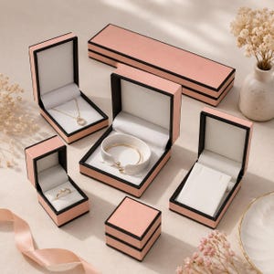 May include: A collection of blush pink jewellery boxes with black trim and white velvet interiors. The set includes a long rectangular box, a square ring box with a gold ring, a necklace box with a gold chain, a bracelet box with a gold bangle, and an empty earring box.