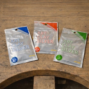 May include: Three silver-colored foil packets labeled ART CLAY SILVER. The left packet has a blue accent and 10g label, the center has an orange accent and 20g label, and the right has a green accent and 7g label. Each packet features a circular hole at the top.