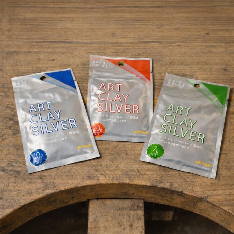 May include: Three silver-colored foil packets labeled ART CLAY SILVER. The left packet has a blue accent and 10g label, the center has an orange accent and 20g label, and the right has a green accent and 7g label. Each packet features a circular hole at the top.