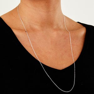 May include: A long, delicate silver chain necklace. The necklace is draped around the neck and hangs down, creating a simple yet elegant look. The chain is thin and appears to be made of a shiny metal.