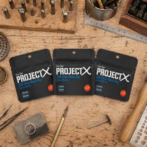 May include: Three black pouches of Five Star PROJECT X .999 Silver Metal Clay, for torch or kiln firing. The pouches contain 5, 15, and 50 grams of clay. The image is set on a wooden surface with jewelry making tools.