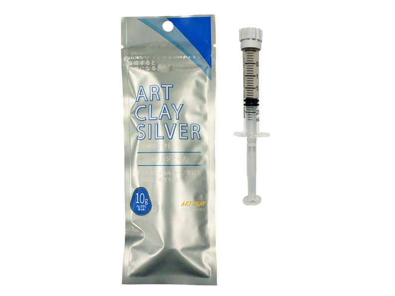 May include: A silver-colored foil pouch labeled ART CLAY SILVER with a 10g weight indicator and a clear plastic syringe containing a gray paste. The syringe features a white plunger and a graduated scale printed on the side of the barrel.