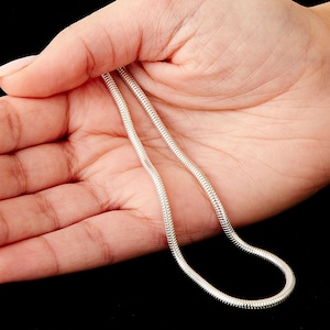 May include: A silver snake chain necklace, held in a hand. The chain has a smooth, rounded profile and a reflective surface, appearing bright against the dark background. The necklace is a classic jewellery piece.