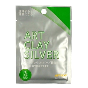 May include: A silver-colored rectangular packet of Art Clay Silver with a green diagonal corner accent. The text reads Art Clay Silver, 7g, Ag 92%, and Japanese characters. It features a circular hole at the top for hanging and a green circular badge.