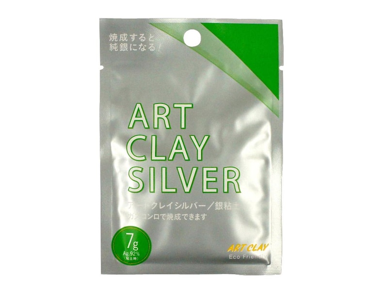 May include: A silver-colored rectangular packet of Art Clay Silver with a green diagonal corner accent. The text reads Art Clay Silver, 7g, Ag 92%, and Japanese characters. It features a circular hole at the top for hanging and a green circular badge.