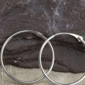 May include: A pair of silver hoop earrings. The earrings are circular and have a simple, elegant design. The hoops are shown against a dark, textured background.