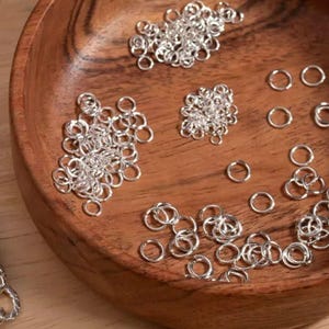 May include: A wooden bowl containing numerous small, shiny silver jump rings. The rings are arranged in small clusters and individually, indicating their use in jewellery making or crafts. The bowl's warm wood grain contrasts with the cool metal.