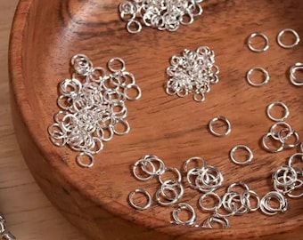 May include: A wooden bowl containing numerous small, shiny silver jump rings. The rings are arranged in small clusters and individually, indicating their use in jewellery making or crafts. The bowl's warm wood grain contrasts with the cool metal.