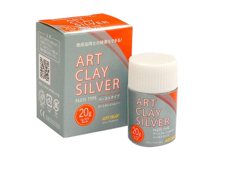 May include: A 20g container of Art Clay Silver paste type and its matching rectangular cardboard box. The packaging is grey and orange with white text that reads Art Clay Silver, Paste Type, and 20g. The container has a white screw-on lid.