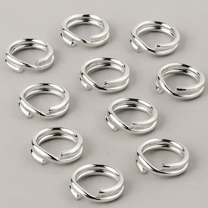 May include: Nine silver-coloured double-loop jump rings are displayed. Each ring features two parallel metal bands, with a small, rounded extension. The rings are arranged on a light grey surface.