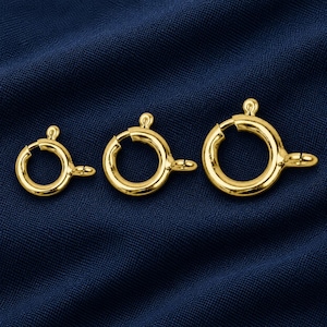May include: Three gold-toned spring ring clasps arranged in a row from smallest to largest. Each circular jewellery finding features a small spring-loaded lever, a tiny protruding knob for operation, and a fixed loop for attachment to a chain or cord.