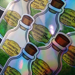 May include: A close-up of several iridescent holographic stickers featuring a cartoon illustration of a glass bottle with a cork stopper. The bottle label reads "Dehydrated Gelatinous Cube, Just Add Water".