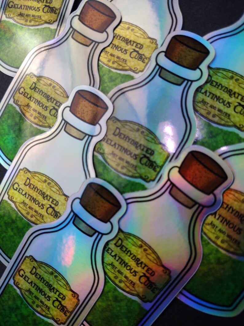 May include: A close-up of several iridescent holographic stickers featuring a cartoon illustration of a glass bottle with a cork stopper. The bottle label reads "Dehydrated Gelatinous Cube, Just Add Water".