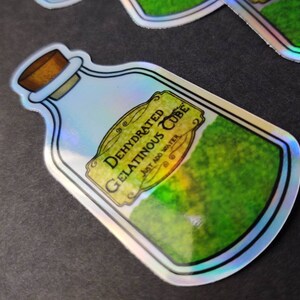 May include: A holographic sticker of a glass bottle containing a green, mossy substance. The label reads "Dehydrated Gelatinous Cube, Just Add Water".