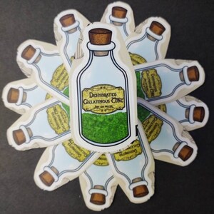 May include: A sticker of a glass bottle with a cork stopper. The bottle is filled with a green substance and has a yellow label that reads "Dehydrated Gelatinous Cube. Just add water."