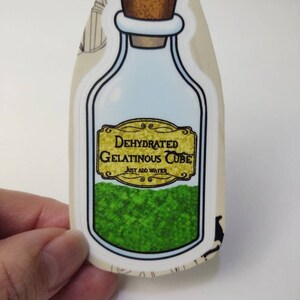May include: A sticker of a glass bottle with a cork stopper. The bottle contains a green powder and has a label that reads "Dehydrated Gelatinous Cube, Just Add Water".