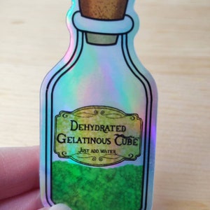 May include: A holographic sticker of a glass bottle with a cork stopper. The bottle is filled with a green substance and has a label that reads "Dehydrated Gelatinous Cube" and "Just Add Water".