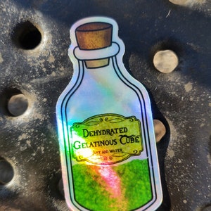 May include: A holographic sticker of a glass bottle with a cork stopper. The bottle is filled with a green liquid and has a label that reads "Dehydrated Gelatinous Cube, Just Add Water".