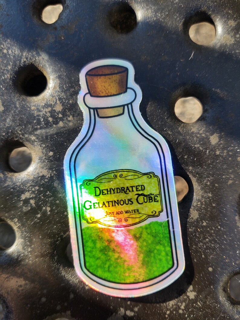 May include: A holographic sticker of a glass bottle with a cork stopper. The bottle is filled with a green liquid and has a label that reads "Dehydrated Gelatinous Cube, Just Add Water".