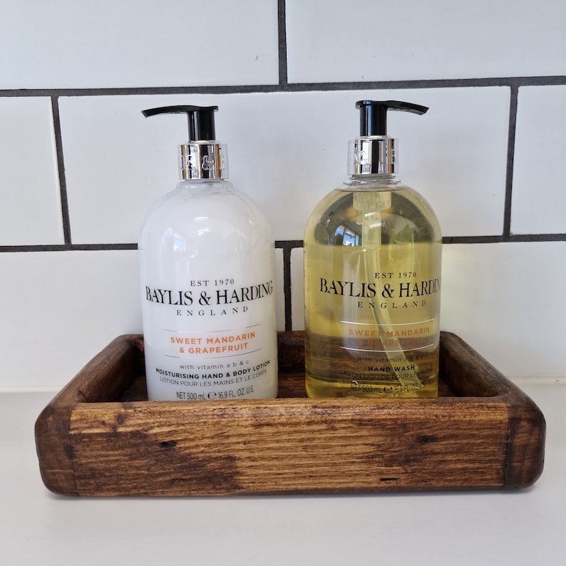 May include: A wooden tray holds two bottles of Baylis & Harding Sweet Mandarin & Grapefruit lotion and hand wash. The bottles have black pumps and silver accents. The lotion bottle is white, and the hand wash is yellow. The tray is made of dark wood.