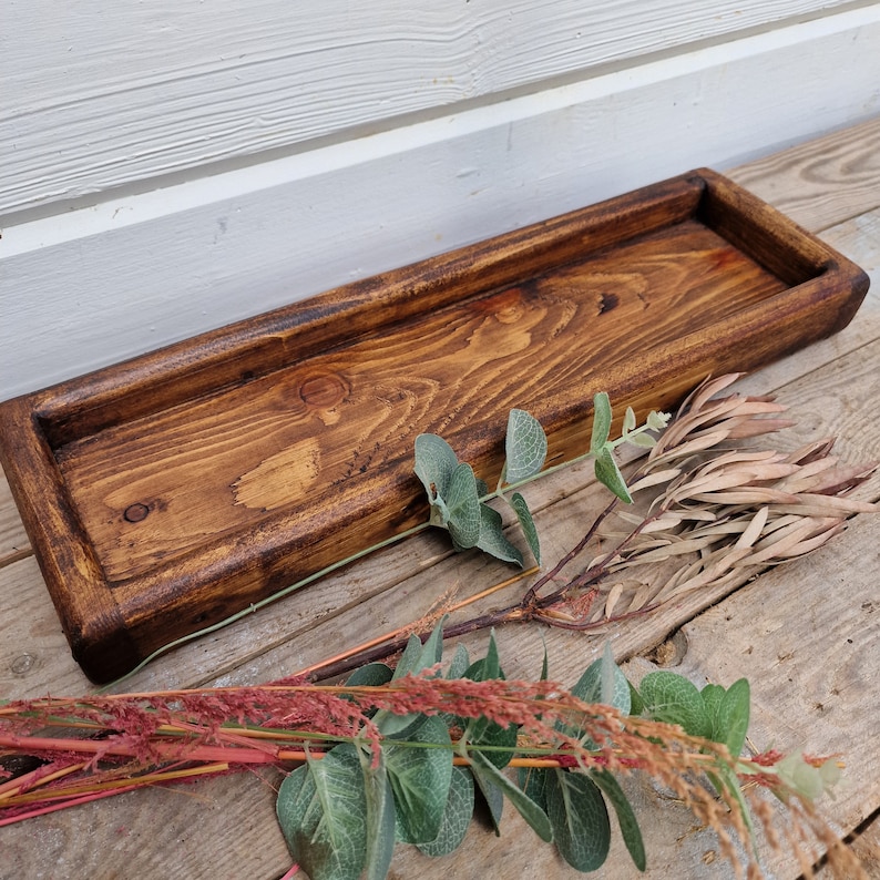 May include: A rectangular, wooden serving tray with a dark brown stain. The tray has raised edges and a recessed interior. Decorative greenery, including eucalyptus and dried flowers, is arranged around the tray. The wood grain is visible.