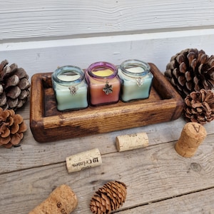 May include: Three square glass candles in pastel colors sit on a wooden tray. Each candle has a metal charm and is surrounded by pine cones and corks. The candles are in shades of green, pink, and yellow.