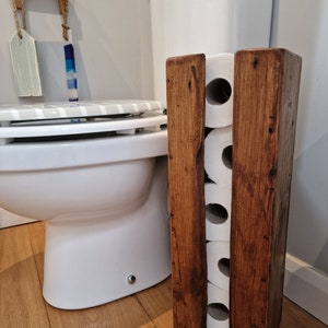 May include: A rustic wooden toilet paper holder with four slots for rolls. The holder is made of dark brown wood and has a natural finish. It is standing next to a white toilet in a bathroom.
