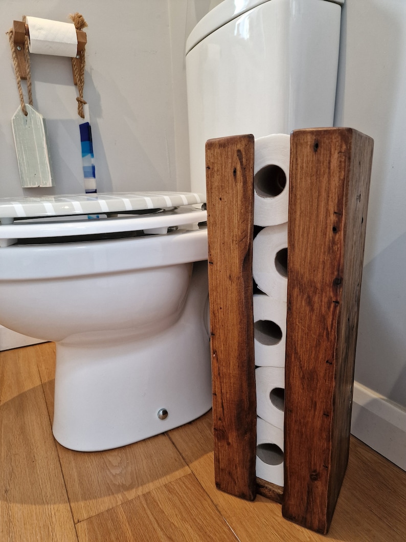 May include: A rustic wooden toilet paper holder with four slots for rolls. The holder is made of dark brown wood and has a natural finish. It is standing next to a white toilet in a bathroom.