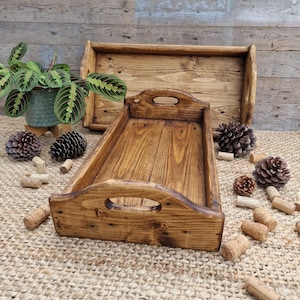 May include: Two rectangular wooden trays with a dark brown stained finish and cutout handles on the ends. The trays feature visible wood grain and are arranged with a potted green patterned plant, several pinecones, and scattered corks.