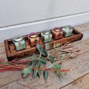 May include: Five square glass jar candles in a wooden tray. Each candle has a different color wax and a butterfly charm. The tray is dark brown, and the candles are arranged on a wooden surface with greenery.