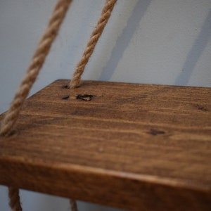 K&ouml;nnte beinhalten: Ein Holzregal mit einer dunkelbraunen Beize, das an zwei dicken Seilen h&auml;ngt. Das Regal hat einen rustikalen Look und ist perfekt f&uuml;r die Pr&auml;sentation kleiner Gegenst&auml;nde.