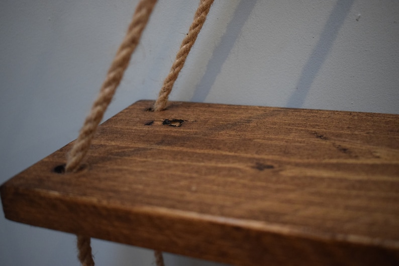 K&ouml;nnte beinhalten: Ein Holzregal mit einer dunkelbraunen Beize, das an zwei dicken Seilen h&auml;ngt. Das Regal hat einen rustikalen Look und ist perfekt f&uuml;r die Pr&auml;sentation kleiner Gegenst&auml;nde.