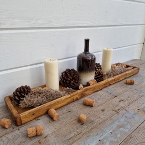 May include: A rustic wooden tray with a brown and white ceramic bottle, two white candles, and pine cones. The tray is on a wooden surface with cork stoppers scattered around.