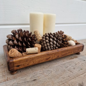 May include: A wooden tray, stained a warm brown, holding pine cones, wine corks, and two ivory pillar candles. The tray sits on small, rounded feet. The arrangement creates a natural, cosy aesthetic, perfect for a home setting.