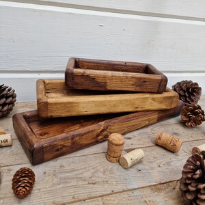 May include: Three handcrafted wooden trays of varying sizes, stained in shades of brown. The trays have raised edges and a rectangular shape. Corks and pinecones are scattered around the trays, suggesting a rustic theme.