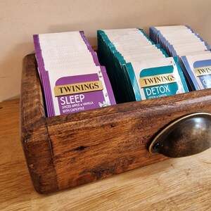 May include: A wooden tea bag organizer with a metal handle. The organizer is filled with boxes of Twinings tea bags. The tea flavors are Sleep, Spiced Apple & Vanilla with Camomile, Detox, Lemon & Ginger.